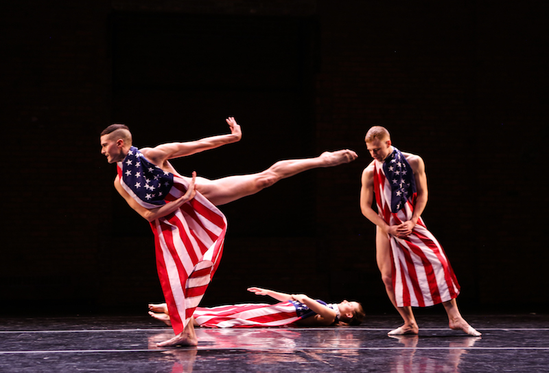 Three dancers where American Flags tied around their necks. One dancer executes a high arabesque. Another lies prone on the floor while the third leans on his right side.